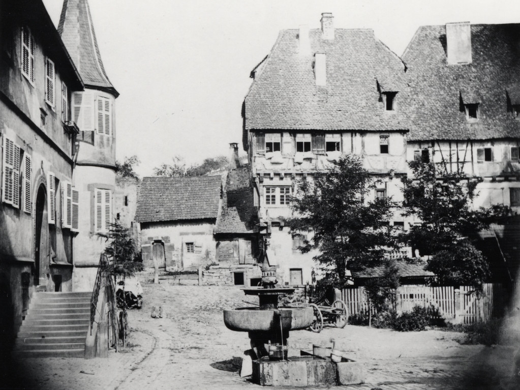Schwarz-weiß Foto von 1890, das ein Ensemble von Gebäuden zeigt, darunter ein Jagdschloss, in der Mitte ein Brunnen